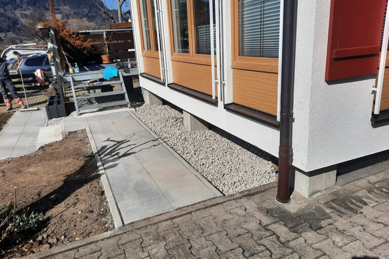 Ein modernes Gebäude mit Holzrahmenfenstern steht auf einem Steinfundament. Entlang des Gebäudes verläuft ein gepflasterter Weg, der von Kies und einem kleinen Gartenbereich mit Tageslicht gesäumt wird. Die Gärtner im Allgäu