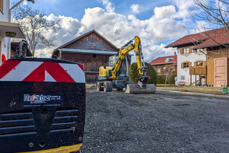 Ein gelber Bagger arbeitet auf einer Schotterstraße in einem Wohngebiet mit Holzhäusern und wolkenverhangenem Himmel; im Vordergrund sind rot-weiß gestreifte Baumaschinen zu sehen. Die Gärtner im Allgäu