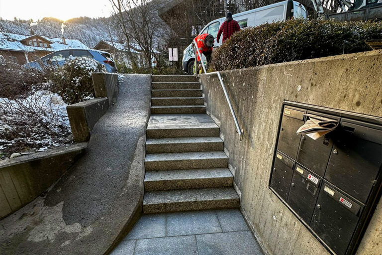 Ein Arbeiter in einer orangefarbenen Weste schaufelt Schnee von den Stufen neben einem abschüssigen Weg mit einem Geländer. In der Nähe befinden sich Briefkästen an einer Betonwand und ein weißer Lieferwagen; im Hintergrund sind Häuser und verschneite Hügel zu sehen. Die Gärtner im Allgäu
