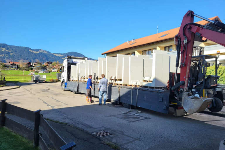 Ein mit großen weißen Schränken beladener Pritschenwagen ist auf einer Straße in der Nähe eines gelben Gebäudes geparkt. Zwei Personen stehen neben dem Lkw und unterhalten sich, und am Heck ist ein Kranarm angebracht. Im Hintergrund sind Berge und Grünflächen zu sehen. Die Gärtner im Allgäu