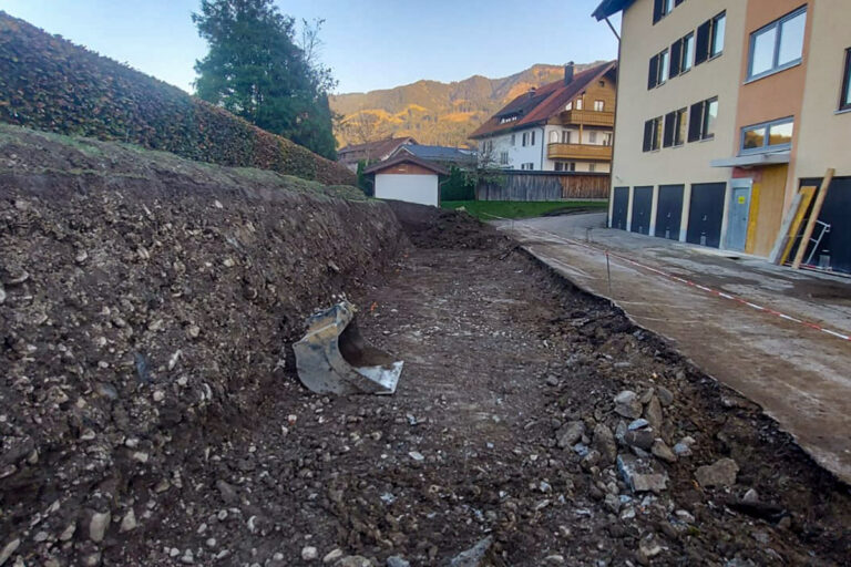 Eine Baustelle neben Wohngebäuden zeigt einen ausgehobenen Bereich mit freiliegender Erde und Steinen, der von einer Hecke und einer kleinen Garage im Hintergrund begrenzt wird. Die Gärtner im Allgäu