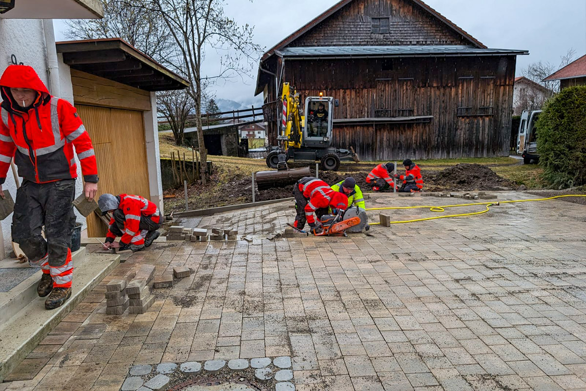 An einem bewölkten Tag verlegen mehrere Arbeiter in roter und orangefarbener Schutzkleidung Pflastersteine und bedienen Maschinen auf einer Baustelle im Freien in der Nähe einer Holzscheune. Die Gärtner im Allgäu