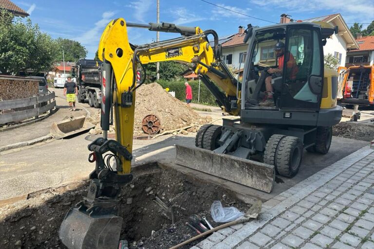 Ein kleiner gelber Bagger gräbt ein Loch in einer Wohnstraße, während ein Arbeiter in der Kabine sitzt. Im Hintergrund sind Erdhaufen, Baumaterialien und andere Arbeiter zu sehen. Die Gärtner im Allgäu
