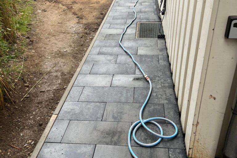 Ein hellblauer Gartenschlauch liegt aufgerollt und gespannt auf einem gepflasterten Gehweg neben einem weißen Gebäude, wobei auf der linken Seite etwas Schmutz und Gras zu sehen sind. Die Gärtner im Allgäu