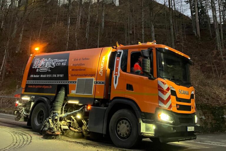 Ein leuchtend orangefarbener Kehrwagen mit SCANIA-Schriftzug steht in der Abenddämmerung an einer Straße in der Nähe eines bewaldeten Hangs. Auf dem Fahrersitz ist eine Person mit einer orangefarbenen Jacke zu sehen. Die Gärtner im Allgäu