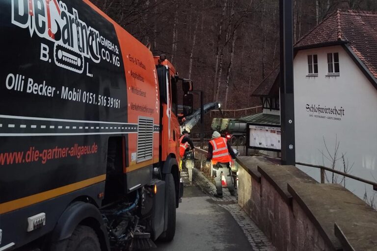 Arbeiter in orangefarbenen Sicherheitswesten bedienen Straßeninstandhaltungsgeräte auf einer schmalen Brücke neben einem Gebäude, während in der Nähe ein Service-Lkw geparkt ist und im Hintergrund bewaldete Hügel zu sehen sind. Die Gärtner im Allgäu