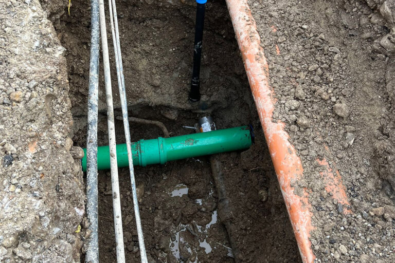 In einem schlammigen Graben liegt ein grünes unterirdisches Rohr frei, daneben befinden sich mehrere Kabel und ein schwarzes vertikales Rohr. Das umliegende Erdreich wurde aufgegraben, um die Infrastruktur unter der Oberfläche freizulegen. Die Gärtner im Allgäu