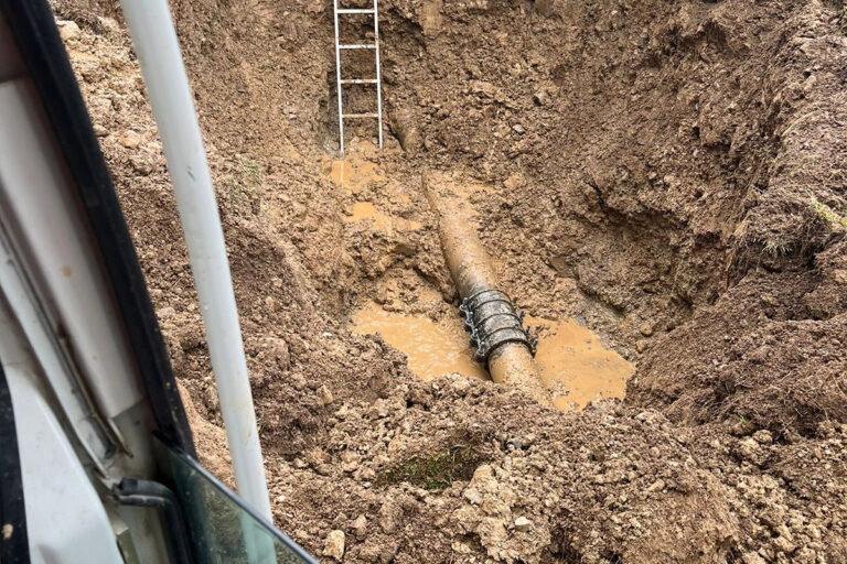 Ein schlammiger Graben, durch den ein großes freiliegendes Rohr verläuft, das teilweise mit Wasser und Schlamm bedeckt ist. Eine Metallleiter lehnt an der Grabenwand, und im linken Vordergrund ist ein Teil eines Fahrzeugs zu sehen. Die Gärtner im Allgäu