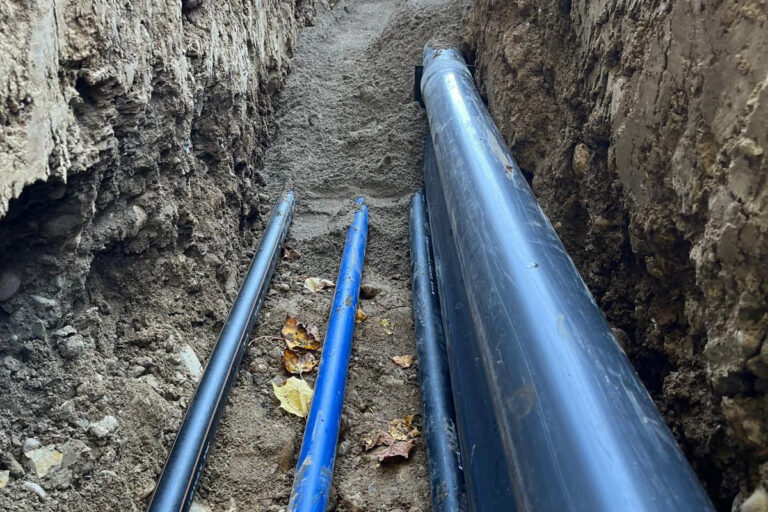 Mehrere große schwarze und blaue Rohre liegen in einem in die Erde gegrabenen Graben, umgeben von Erde und ein paar verstreuten Blättern. Die Rohre verlaufen parallel und scheinen Teil einer unterirdischen Anlage zu sein. Die Gärtner im Allgäu