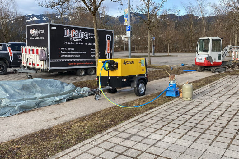 Ein gelb-schwarzer mobiler Luftkompressor ist über einen Schlauch mit einem Propantank und einer Gasflasche auf einem Parkplatz verbunden. In der Nähe stehen ein Anhänger, ein kleiner Bagger und einige Baumaterialien. Im Hintergrund sind Bäume und Berge zu sehen. Die Gärtner im Allgäu