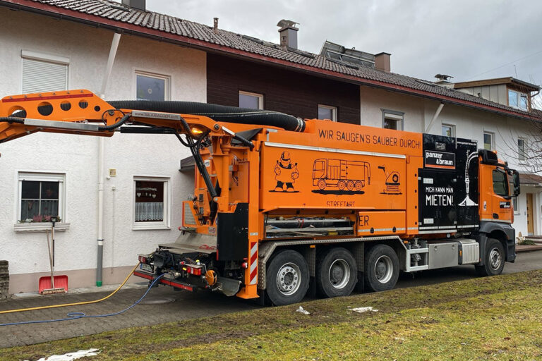 Ein orangefarbener Industriestaubsauger mit einem großen Schlauch ist neben einem weißen Gebäude geparkt und fährt den Schlauch zum Reinigen durch ein Fenster aus. Der Lkw hat deutsche Texte und Illustrationen auf seiner Seite. Die Gärtner im Allgäu