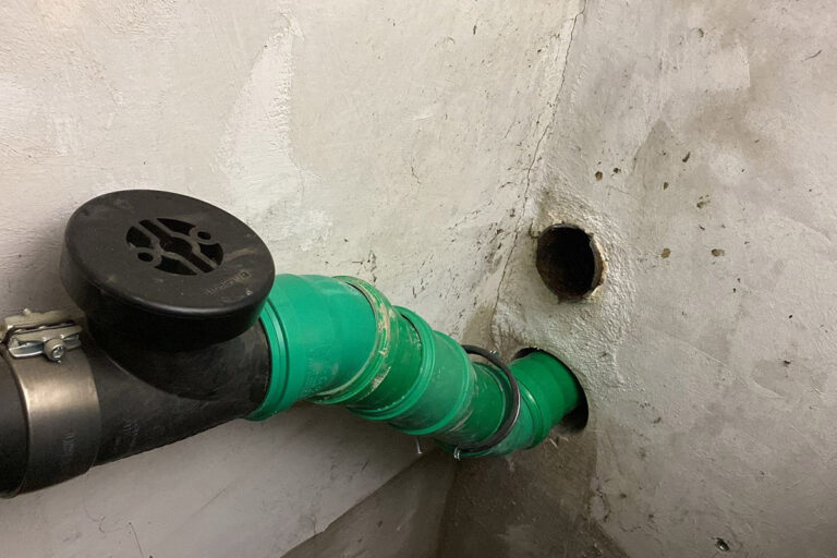 Ein grünes Rohr mit schwarzen Anschlüssen mündet in ein Loch in einer weißen, unfertigen Wand. Daneben befindet sich ein weiteres leeres Loch ähnlicher Größe in der Wand. Der Bereich scheint Teil eines Kellers oder Versorgungsraums zu sein. Die Gärtner im Allgäu