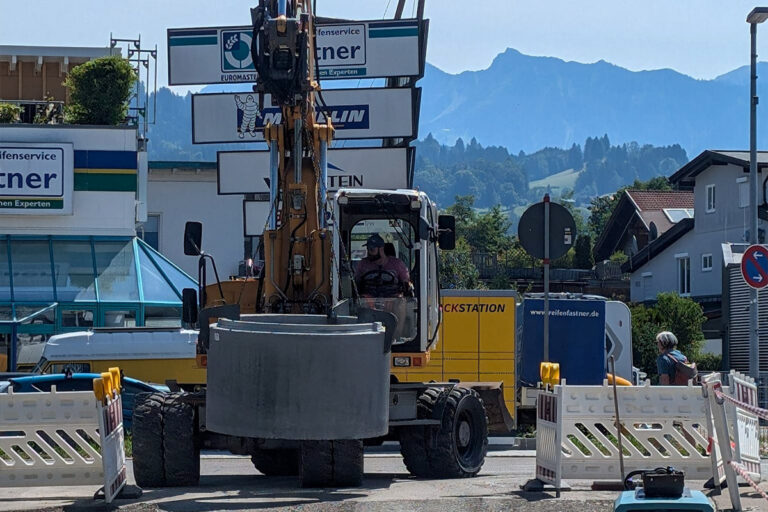 Ein Bagger blockiert eine Straße, umgeben von weißen Schranken und Schildern. Eine Person bedient die Maschine, im Hintergrund sind an einem sonnigen Tag Gebäude und Berge zu sehen. Die Gärtner im Allgäu