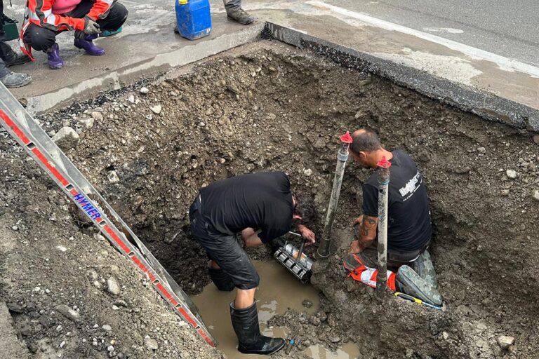 Zwei Arbeiter reparieren in einer schlammigen Ausgrabungsstätte Geräte in einem mit Wasser gefüllten Loch auf einer Straße. Einer kniet in der Nähe des Geräts, während der andere in Stiefeln steht. Werkzeuge und eine weitere Person sind in der Nähe zu sehen. Die Gärtner im Allgäu