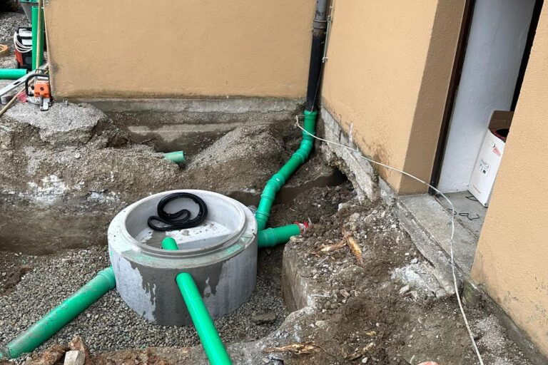 Grüne Kunststoffrohre sind an einen runden Kontrollschacht aus Beton außerhalb eines beigen Gebäudes angeschlossen. Die Baustelle ist von Kies und Erde umgeben, und rechts ist eine Türöffnung zu sehen. Die Gärtner im Allgäu