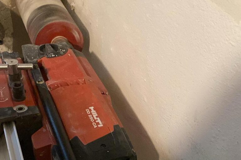 Eine Nahaufnahme einer roten Hilti DD 250-CA Kernbohrmaschine, die an einer beigen Wand montiert ist, wobei die zylindrische Bohrkrone zum Bohren horizontal positioniert ist. Die Gärtner im Allgäu