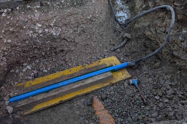 Ein blaues Rohr und ein blaues Kabel liegen auf einer gelben Platte, umgeben von Schotter und Erde auf einer Baustelle. Ein orangefarbenes Rohr und ein blauer Ventilschlüssel sind in der Nähe teilweise sichtbar. Die Gärtner im Allgäu