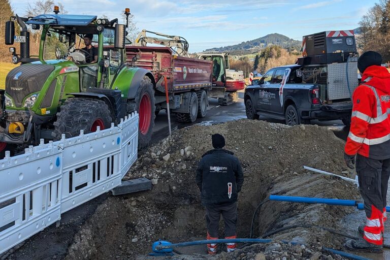 Mehrere Arbeiter in Schutzkleidung befinden sich auf einer Baustelle am Straßenrand mit einem Traktor, einem Kipper und Nutzfahrzeugen. Ein Arbeiter steht in einem Graben neben freiliegenden Rohren, während andere zusehen. Absperrungen trennen die Baustelle vom Verkehr. Die Gärtner im Allgäu