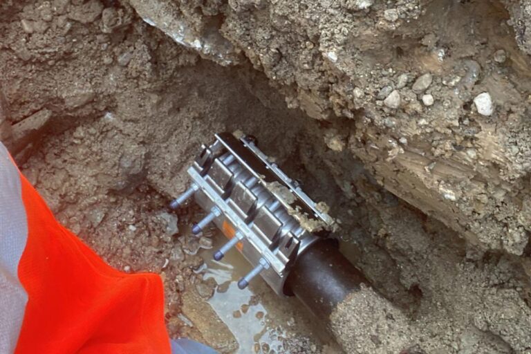 Nahaufnahme einer Metallrohrschelle, mit der ein schwarzes Rohr unter der Erde befestigt wird, umgeben von Schmutz und Steinen. Eine orangefarbene Sicherheitsweste ist teilweise auf der linken Seite des Bildes zu sehen. Die Gärtner im Allgäu