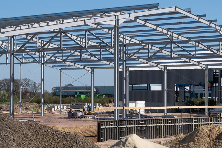 Ein Stahlgerüst eines im Bau befindlichen Gebäudes mit freiliegenden Trägern und Stützen, umgeben von Erdhaufen und Baumaterialien an einem klaren, sonnigen Tag. Die Gärtner im Allgäu