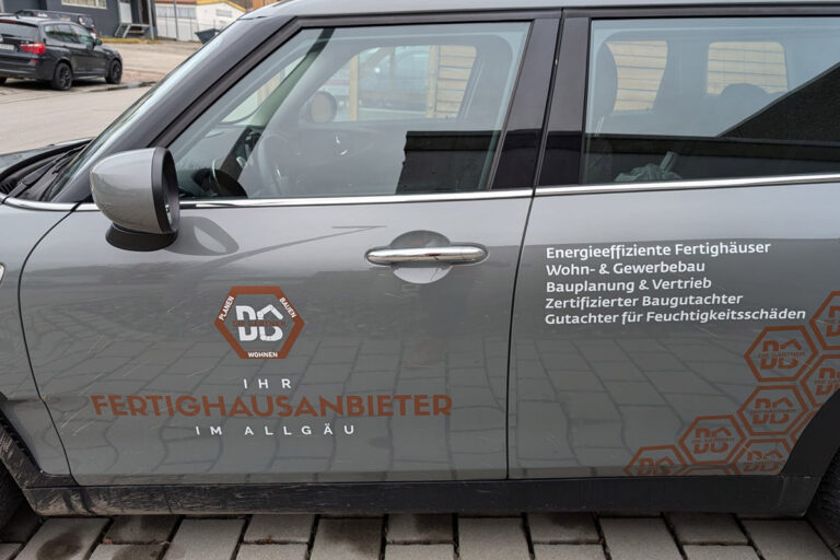 Ein graues Auto mit deutschem Text, das für energieeffiziente Fertighäuser und Baudienstleistungen wirbt, parkt auf einer Steinauffahrt. Das Logo und die angebotenen Dienstleistungen sind an der Fahrertür angebracht. Die Gärtner im Allgäu
