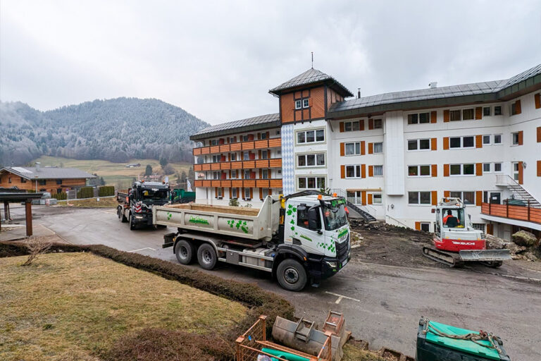 Eine Baustelle vor einem mehrstöckigen Gebäude mit Lastwagen und einem Bagger bei der Arbeit; im Hintergrund sind Berge und frostige Bäume an einem bewölkten Tag zu sehen. Die Gärtner im Allgäu