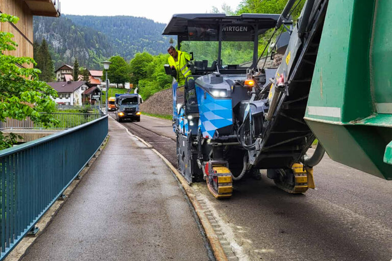 Eine Straßenbaumaschine verlegt neuen Asphalt auf einer Landstraße, die von einem blauen Geländer und Bäumen begrenzt wird. Im Hintergrund sind ein Arbeiter, der die Maschine bedient, und ein weiteres Fahrzeug zu sehen. Die Gärtner im Allgäu