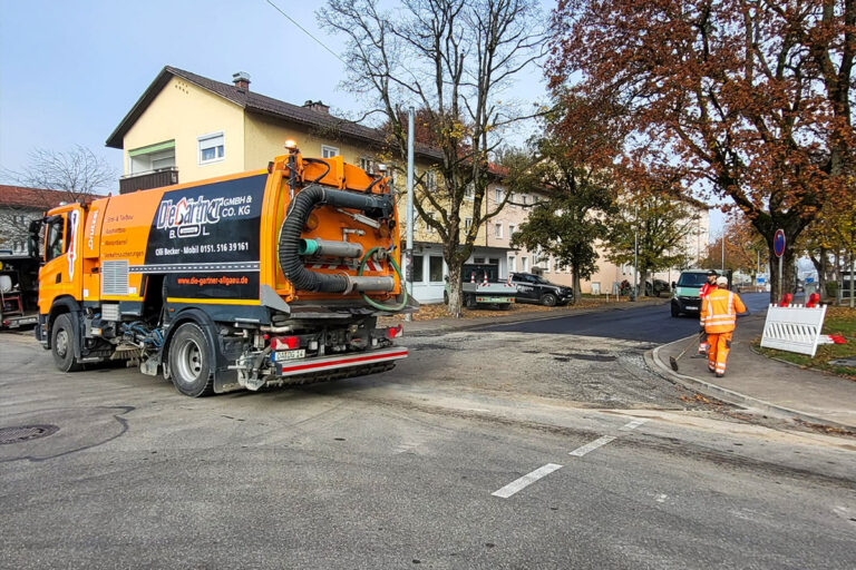 Ein Straßenreinigungsfahrzeug ist am Rande einer Wohnstraße geparkt, während ein Arbeiter in orangefarbener Schutzkleidung auf ihn zugeht. Bäume mit Herbstlaub und mehrere Häuser säumen den Hintergrund. Die Gärtner im Allgäu