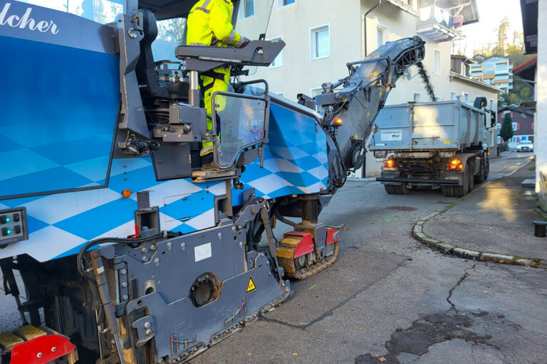 Eine Straßenfräse, die von einem Arbeiter in einem gelben Anzug bedient wird, befördert Asphaltschutt über ein Förderband in einen wartenden Lkw in einer Wohnstraße. Die Gärtner im Allgäu