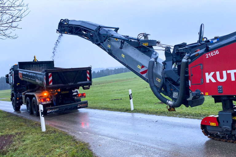 Eine Straßenfräse lädt den gefrästen Asphalt in einen Kipplaster auf einer Landstraße, umgeben von grünen Feldern unter einem bedeckten Himmel. Die Gärtner im Allgäu