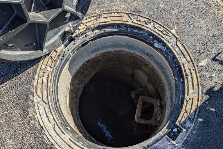 Nahaufnahme eines offenen Schachtes auf Asphalt, dessen runder Metalldeckel teilweise zur Seite gehoben ist. Bei dieser Szene, die typisch für Straßensanierungs- oder Asphaltarbeiten ist, sind Betonwände und einige Ziegelsteine am Boden sichtbar. Die Gärtner im Allgäu