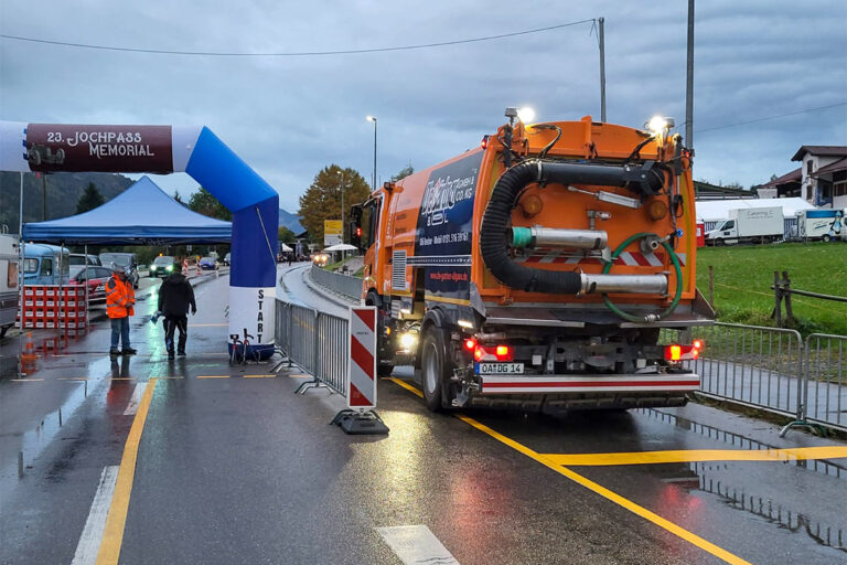 Ein großer orangefarbener Straßenreinigungswagen ist bei einer Veranstaltung im Freien in der Nähe eines blauen Starttors geparkt. Arbeiter in reflektierenden Jacken stehen in der Nähe auf einer nassen Straße, und am Straßenrand sind Absperrungen und Zelte aufgebaut. Die Gärtner im Allgäu