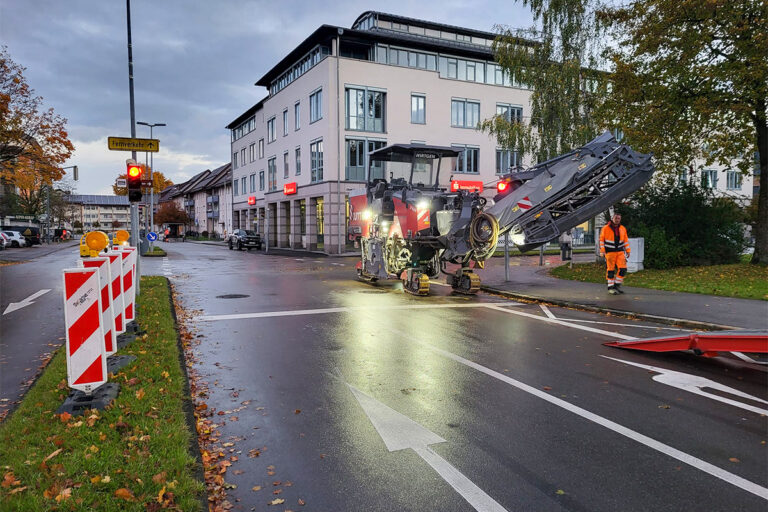 Eine Straßenbauszene zeigt Arbeiter und Maschinen, die in der Morgendämmerung oder in der Abenddämmerung eine Stadtstraße erneuern, wobei beleuchtete Warnschilder und Verkehrssperren die Fahrzeuge von der Arbeitszone fernhalten. Die Gärtner im Allgäu