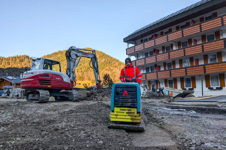 Ein Bauarbeiter in einer leuchtend orangefarbenen Jacke bedient eine Verdichtungsmaschine auf einem unwegsamen, kiesigen Gelände. Im Hintergrund sind ein Bagger und ein mehrstöckiges Gebäude mit Balkonen zu sehen, das von bewaldeten Hügeln umgeben ist. Die Gärtner im Allgäu