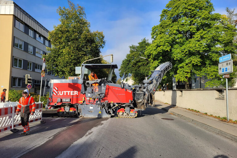 Eine große rote Straßenbaumaschine, die von Arbeitern in orangefarbener Schutzkleidung bedient wird, erneuert eine Straße, während im Hintergrund Bäume, Gebäude und Verkehrsschilder zu sehen sind. Ein Arbeiter steht in der Nähe einer weißen Absperrung. Die Gärtner im Allgäu