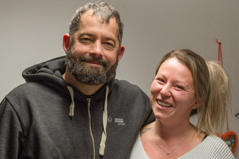 Ein Mann mit Bart und eine Frau mit hellem Haar stehen dicht nebeneinander und lächeln in die Kamera. Der Mann trägt einen dunklen Kapuzenpulli und die Frau einen hellen Pullover. Der Hintergrund ist schlicht und innen. Die Gärtner im Allgäu