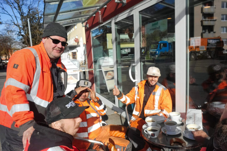Vier Bauarbeiter in orangefarbener, reflektierender Kleidung sitzen und stehen um einen kleinen Cafétisch mit Kaffeetassen und Gebäck, genießen eine Pause und lächeln in die Sonne. Die Gärtner im Allgäu