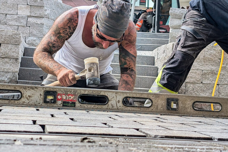 Ein tätowierter Bauarbeiter in einem Tank-Top verwendet einen Hammer und eine Wasserwaage, um Pflastersteine zu verlegen, während ein anderer Arbeiter in der Nähe steht. Im Hintergrund sind Stapel von Steinblöcken und Werkzeugen zu sehen. Die Gärtner im Allgäu