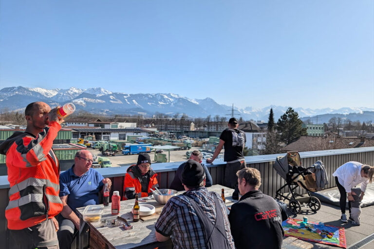Eine Gruppe von Menschen sitzt und steht um einen Tisch auf einer Dachterrasse und teilt Getränke und Essen. Im Hintergrund sind Industriegebäude und schneebedeckte Berge unter einem klaren blauen Himmel zu sehen. Die Gärtner im Allgäu