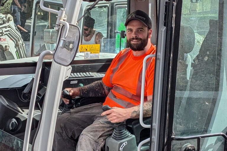 Ein Mann mit einem leuchtend orangefarbenen Sicherheitshemd und einer schwarzen Mütze sitzt in der Kabine einer Baumaschine, lächelt und hält die Bedienelemente. Ein weiterer Arbeiter ist im Hintergrund zu sehen. Die Gärtner im Allgäu