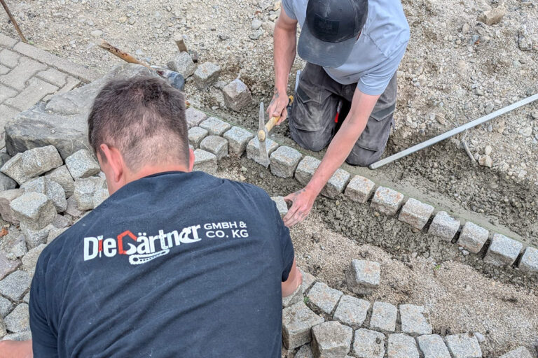 Zwei Arbeiter verlegen Pflastersteine in einem geschwungenen Muster auf einer sandigen Fläche. Ein Arbeiter trägt ein Hemd mit der Aufschrift Die Gärtner GmbH & Co. KG auf dem Rücken; beide sind mit dem Verlegen und Nivellieren der Steine beschäftigt. Die Gärtner im Allgäu