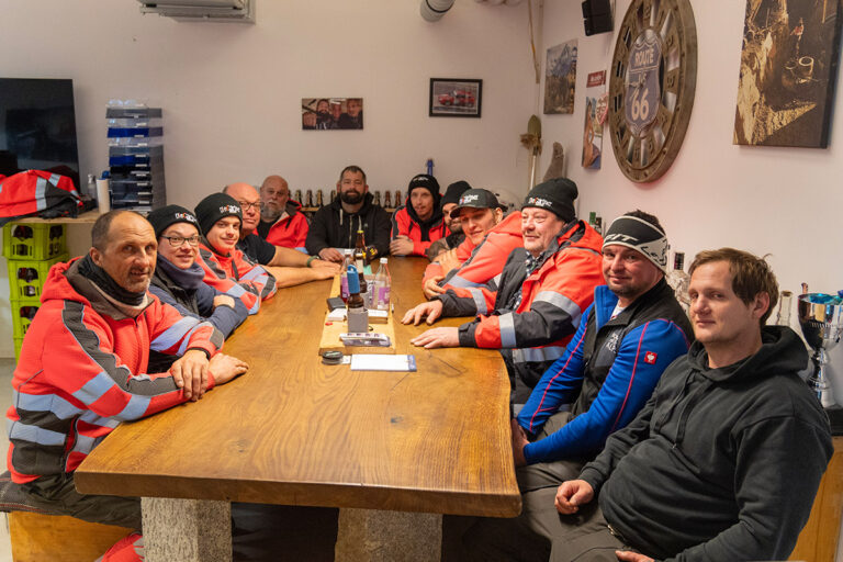 Eine Gruppe von zwölf Personen in Outdoor-Ausrüstung sitzt um einen Holztisch in einem gemütlichen Raum, der mit Erinnerungsstücken und Fotos der Route 66 dekoriert ist, lächelt und schaut in die Kamera. Die Gärtner im Allgäu