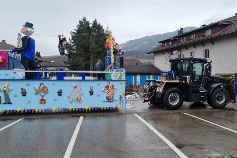 Ein Traktor zieht einen geschmückten Umzugswagen mit Zeichnungen zum Thema Zirkus, darunter Tiere und ein Clown. Menschen in Kostümen stehen auf dem Wagen, und die Szene spielt auf einem Parkplatz an einem bewölkten Tag. Die Gärtner im Allgäu