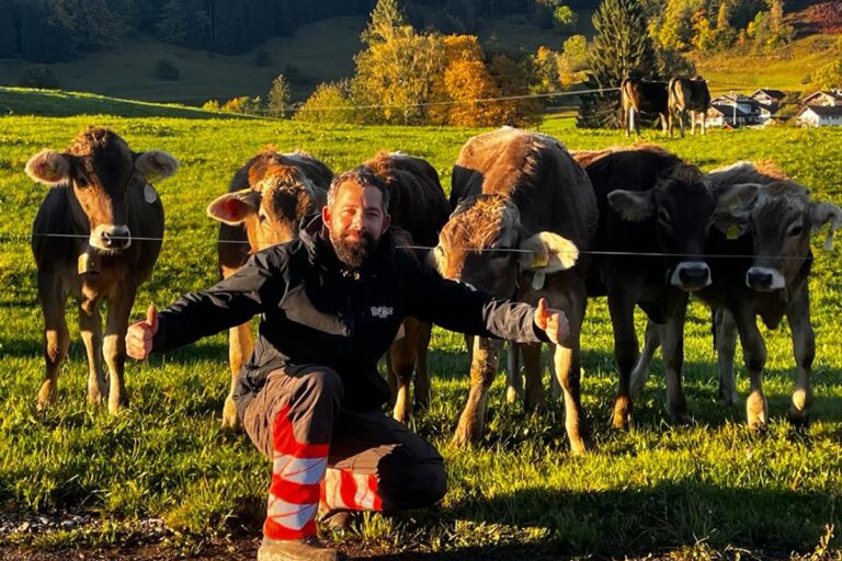Ein Mann kniet vor fünf braunen Kühen auf einer grünen Wiese und lächelt mit erhobenem Daumen. Die Sonne scheint, und im Hintergrund sind Bäume und Hügel zu sehen. Zwei weitere Kühe grasen weiter hinten. Die Gärtner im Allgäu