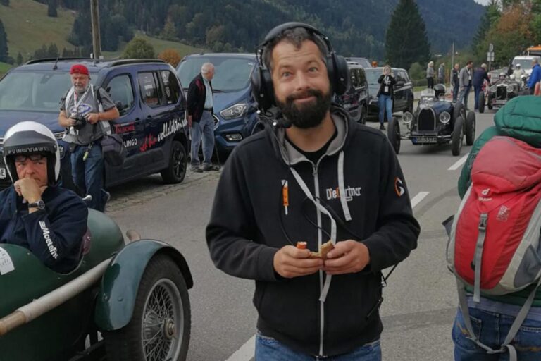 Ein bärtiger Mann mit Kopfhörern und schwarzem Kapuzenpulli steht am Straßenrand und hält einen Imbiss in der Hand, während im Hintergrund Oldtimer und Menschen bei einer Veranstaltung im Freien zu sehen sind. Die Gärtner im Allgäu