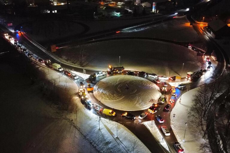 Nächtliche Luftaufnahme eines verschneiten Kreisverkehrs voller Autos und Lastwagen, deren Scheinwerfer die Straße erhellen und einen Verkehrsstau verursachen. Die Umgebung ist leicht verschneit, die Straßen und Gebäude in der Nähe sind schwach beleuchtet. Die Gärtner im Allgäu
