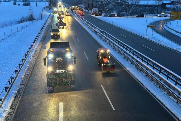 Ein Konvoi von Lastwagen fährt auf einer verschneiten Autobahn. Ein Lkw trägt ein deutsches Schild mit der Aufschrift Unser Geld muss in Deutschland bleiben. Der Boden und die Felder in der Nähe sind mit Schnee bedeckt, und der Himmel ist bedeckt. Die Gärtner im Allgäu