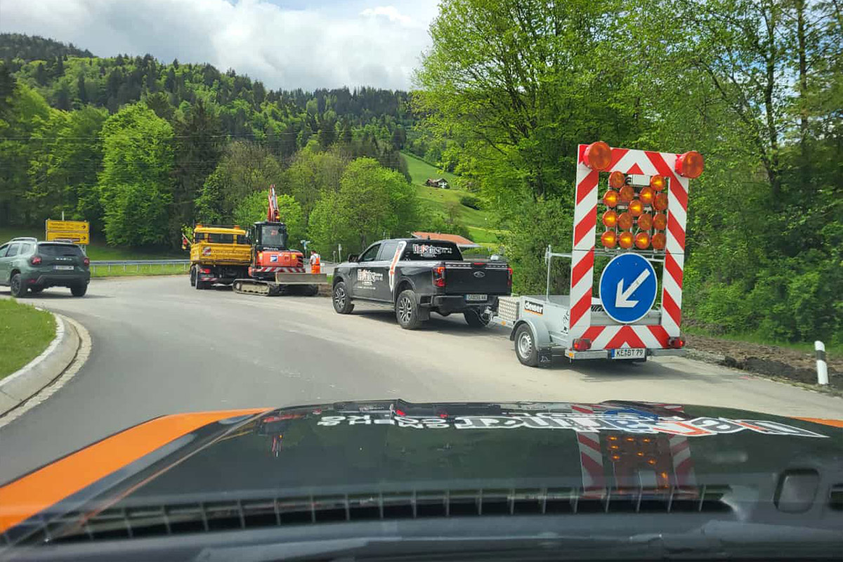 Eine Baustellenszene auf einer Landstraße mit einem großen Pfeilschild, das den Verkehr nach rechts lenkt, mehreren Fahrzeugen, darunter ein Lkw mit Baumaschinen, und grünen Bäumen und Hügeln im Hintergrund. Die Gärtner im Allgäu