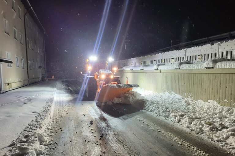 Ein Schneepflug mit hellen Scheinwerfern räumt nachts Schnee von einer schmalen, schneebedeckten Straße, die links von einem Gebäude und rechts von einem Zaun flankiert wird. Am Straßenrand stapelt sich der Schnee. Die Gärtner im Allgäu