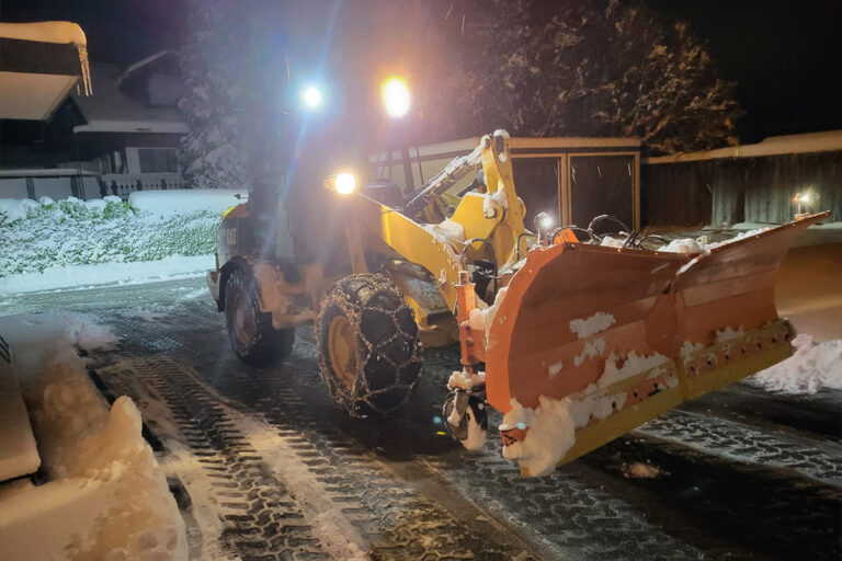 Ein Schneepflug mit eingeschalteten Scheinwerfern räumt bei Nacht eine Einfahrt. Der Schnee bedeckt den Boden, die Bäume und die umliegenden Gebäude, während Reifenspuren den geräumten Bereich markieren. Die Gärtner im Allgäu